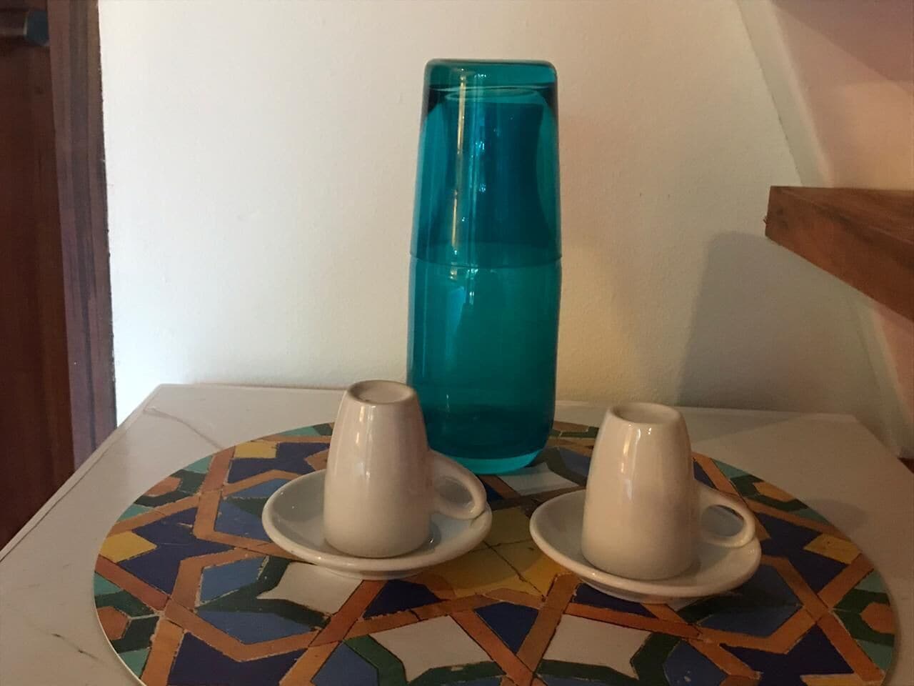 A table with two white cups on saucers and a tall blue vase.