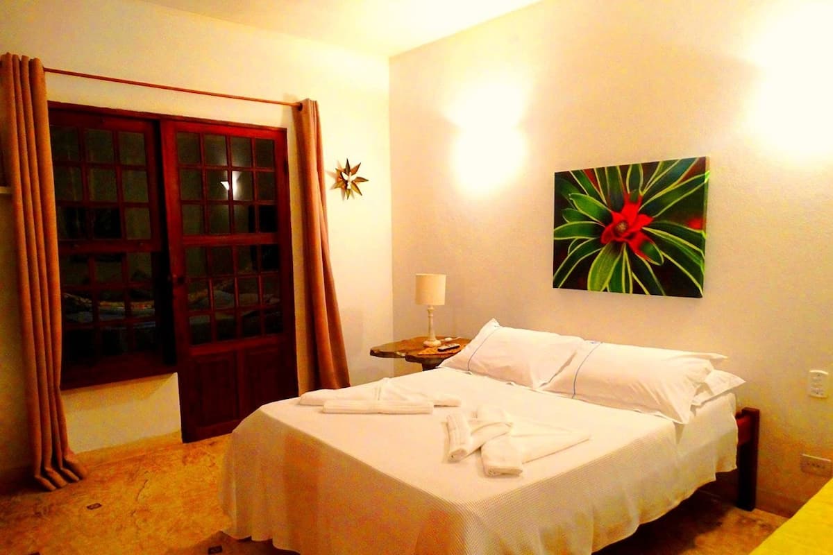 Quarto de hotel com cama, mesa de cabeceira, luminária, janela com cortinas e um objeto decorativo na parede.