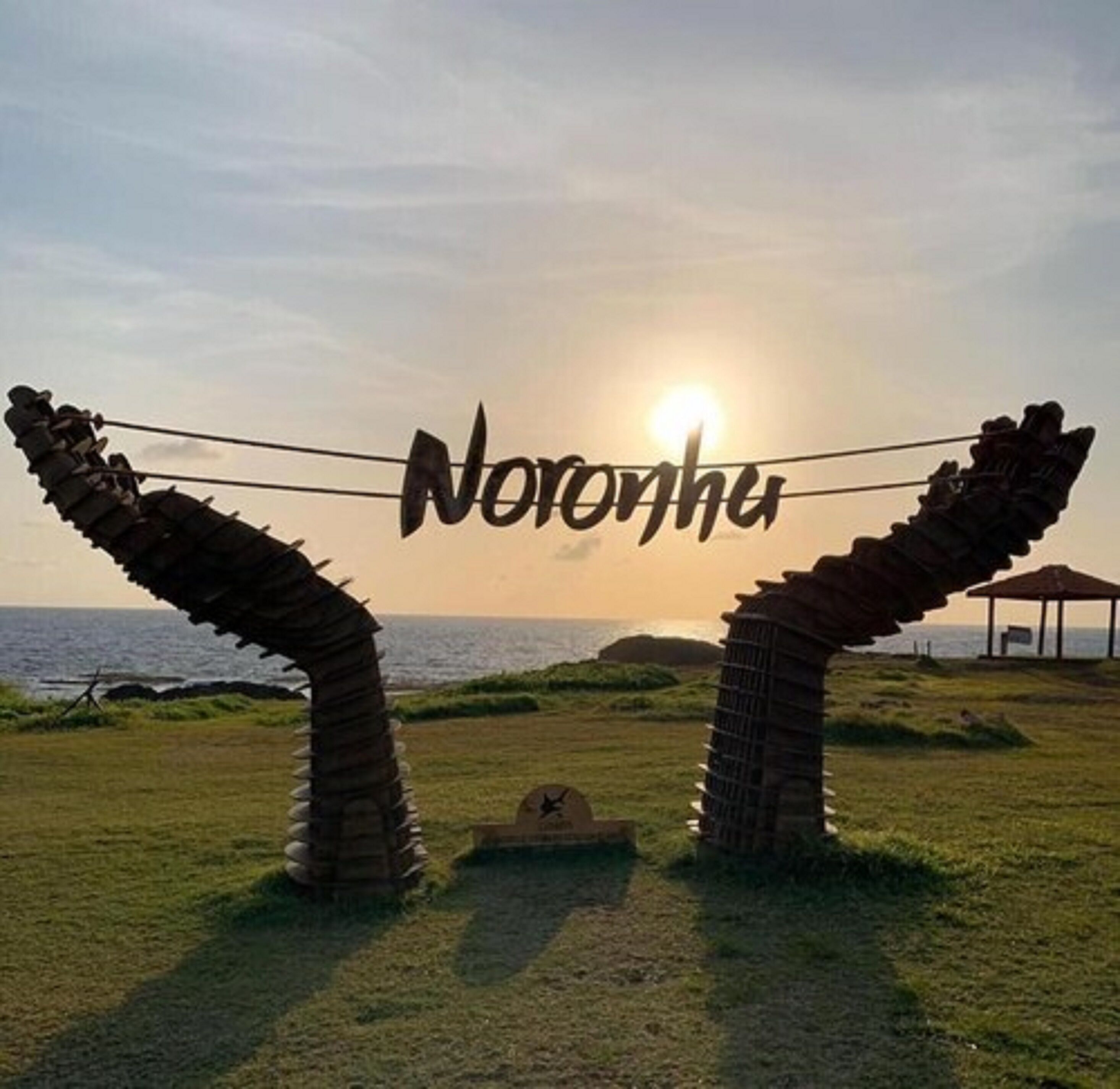 A sunset view with a sign reading "Noronha" and two rock-like structures framing the scene.
