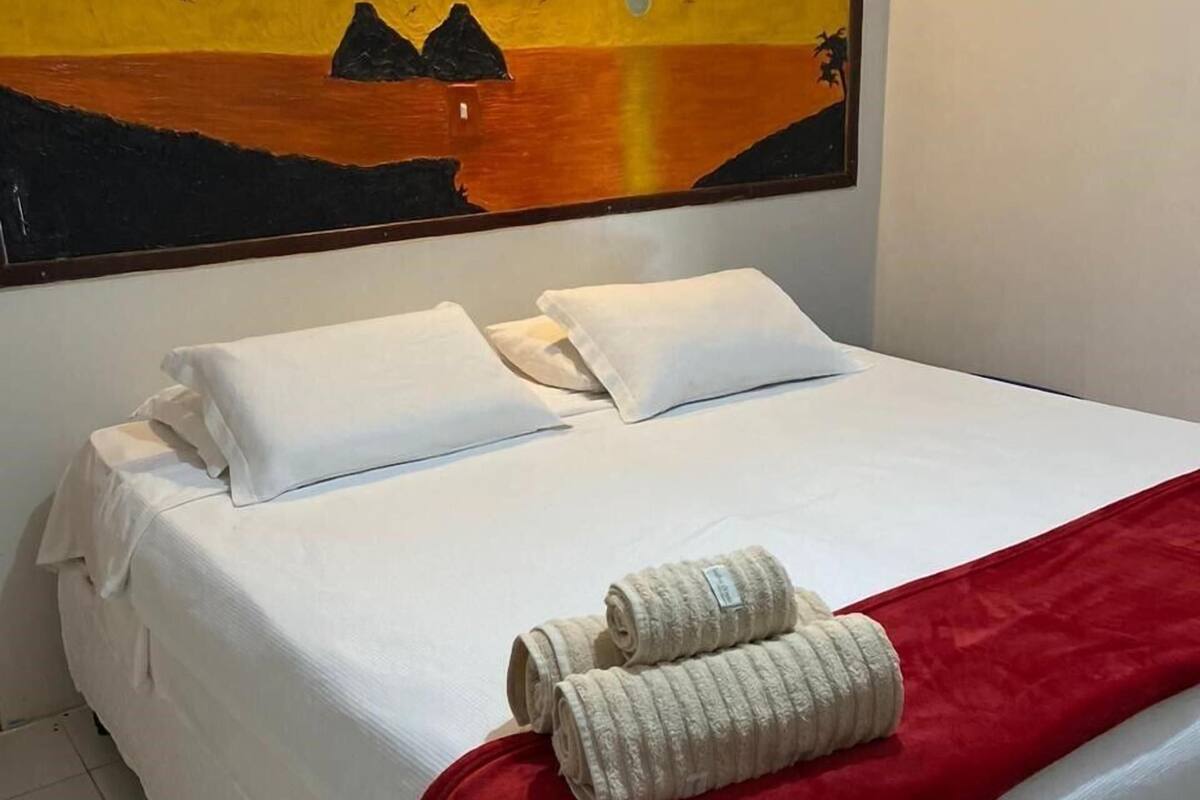 Un lit bien fait, avec des draps blancs, un plaid rouge et deux serviettes enroulées. Un tableau représentant un coucher de soleil sur une étendue d’eau est accroché au mur, au-dessus du lit.