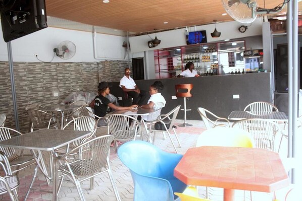 Un café al aire libre con mesas y sillas de metal, una zona de bar al fondo y una pared de piedra.