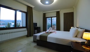Quarto de hotel com uma cama grande, uma escrivaninha com luminária, televisão e vista para a cidade através de amplas janelas.