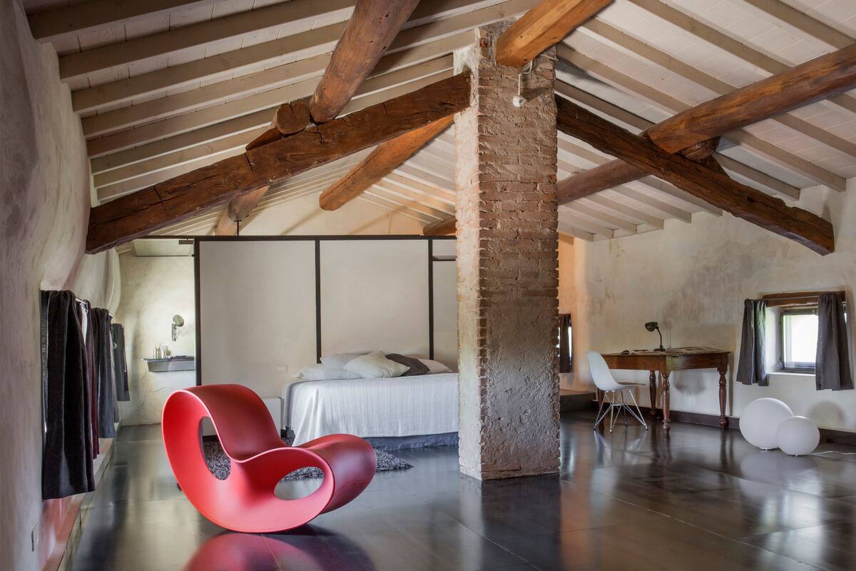 A spacious room with a high ceiling, exposed wooden beams, a bed with white linens, a red chair, and a desk with a chair.