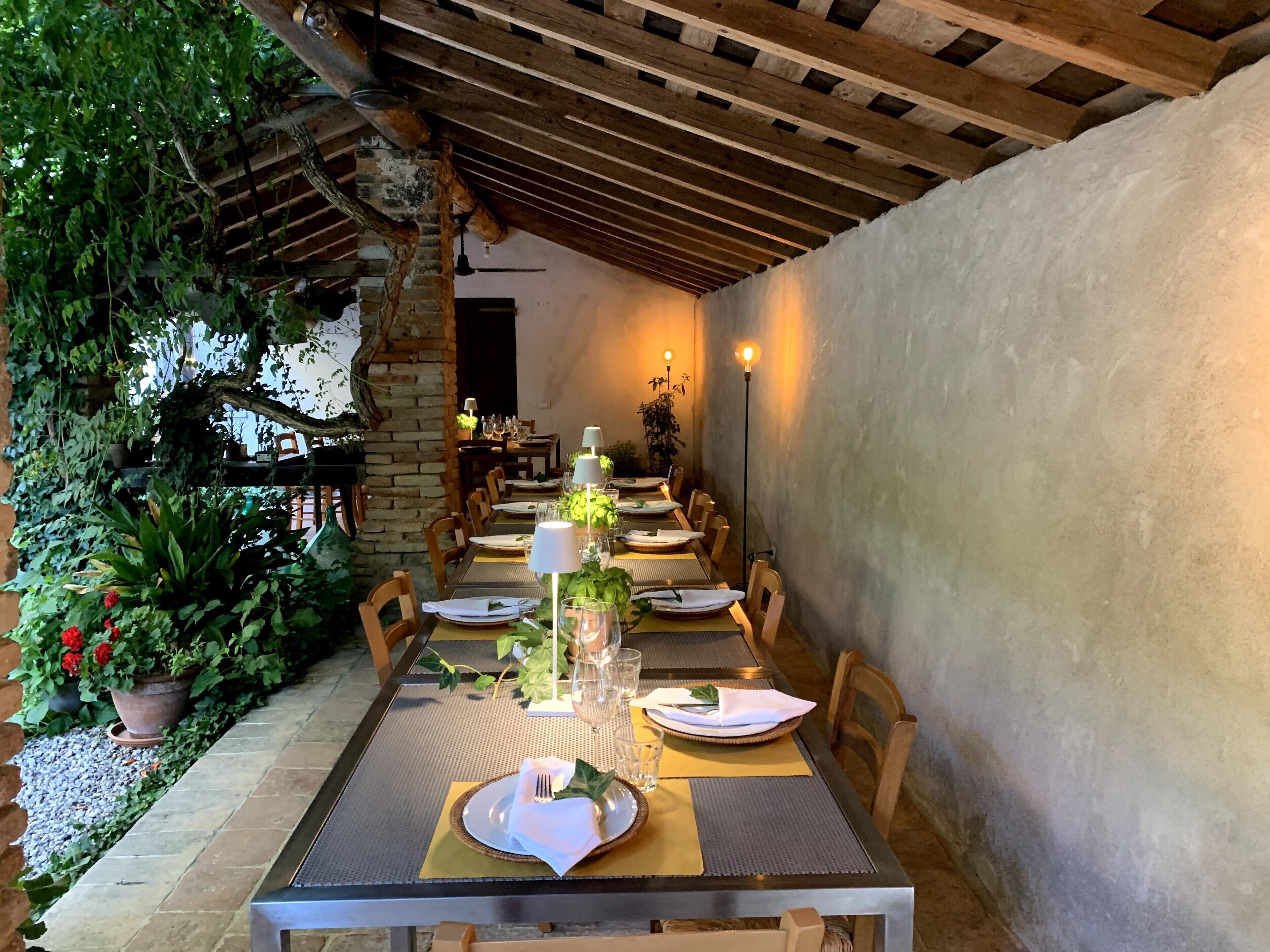 Un área de comedor al aire libre con una larga mesa dispuesta para una comida, rodeada de sillas, bajo una pérgola de madera.