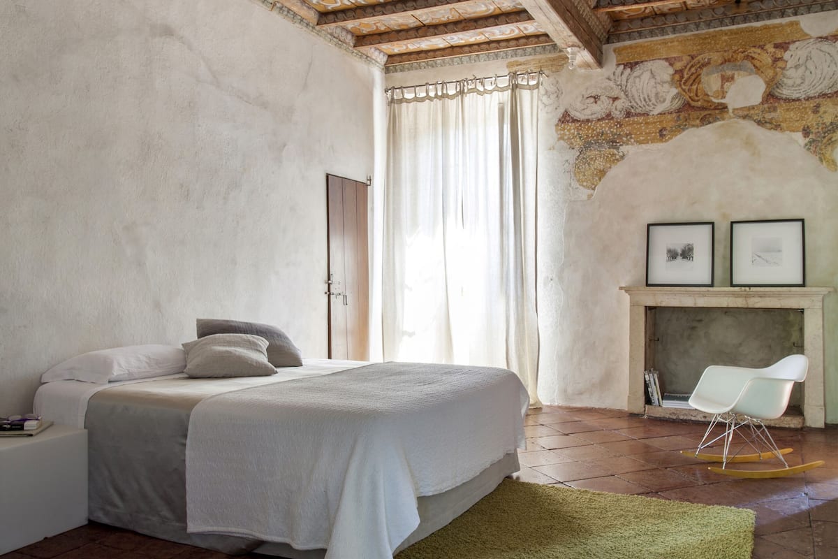 A bedroom with a bed, a green rug, a white chair, and a fireplace.