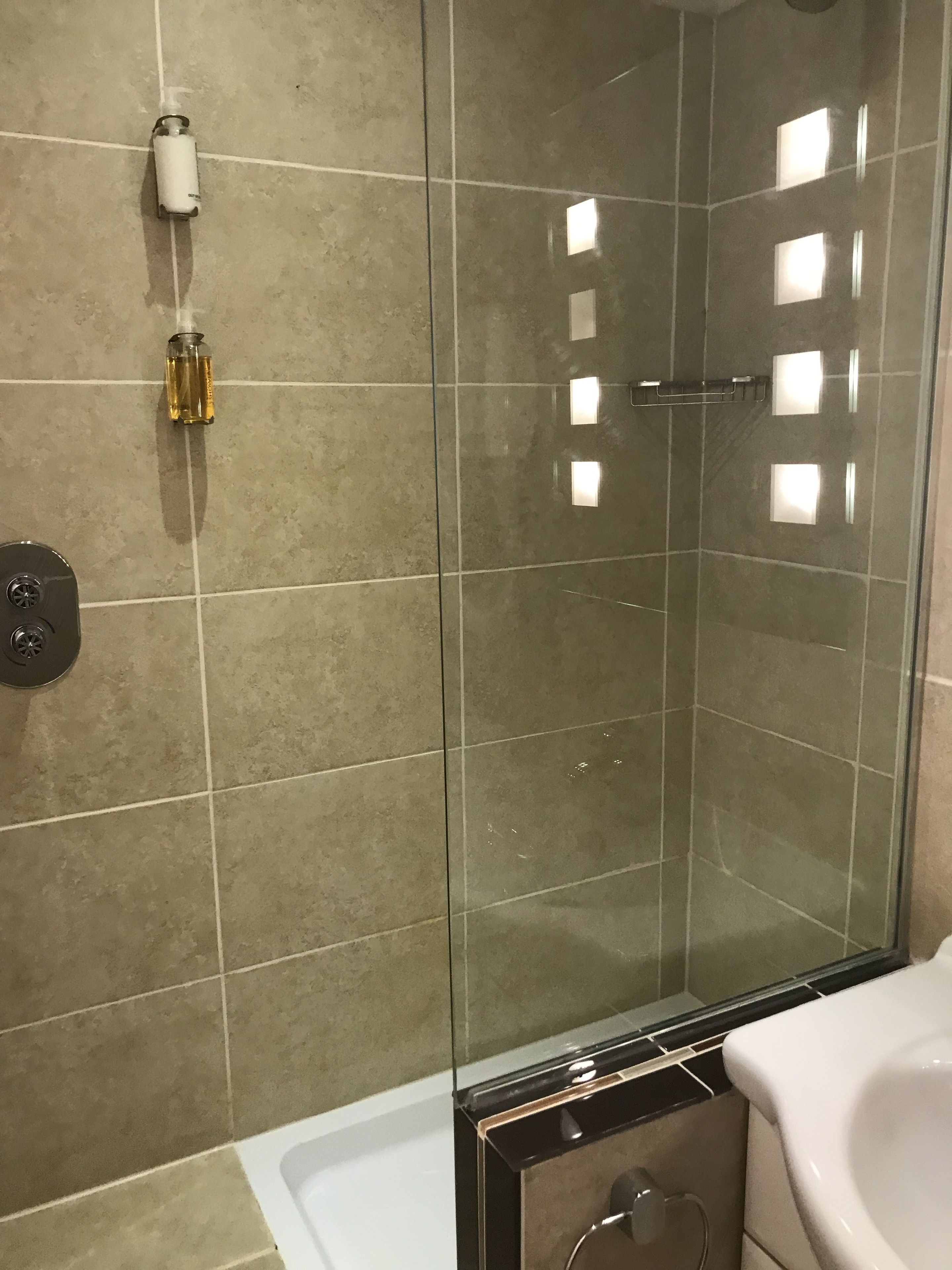 A modern bathroom with a glass-enclosed shower, beige tiles, a white sink, and a soap dispenser on the wall.