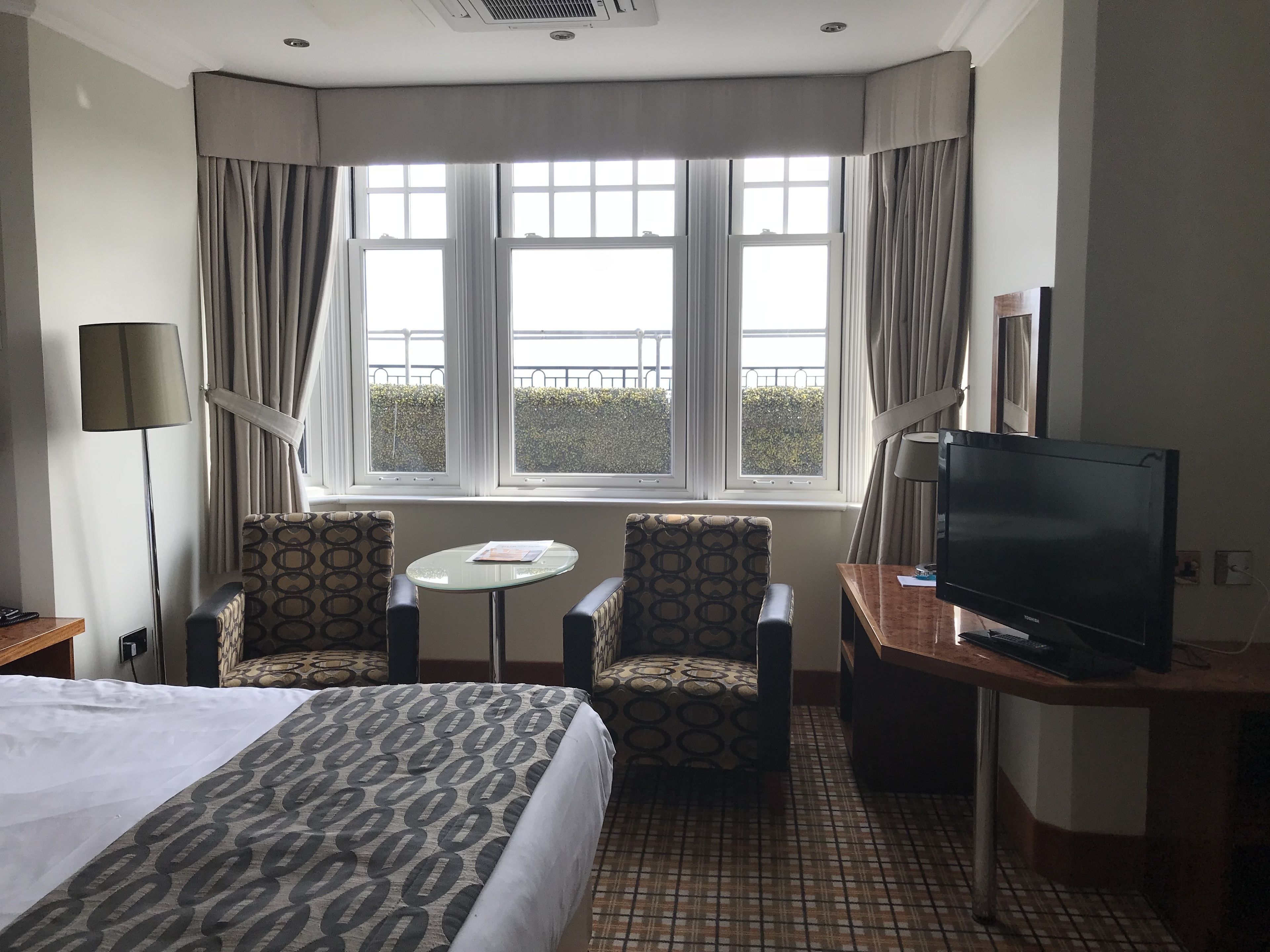 A hotel room with a bed, two patterned armchairs, a small table, a TV, and a window with curtains.