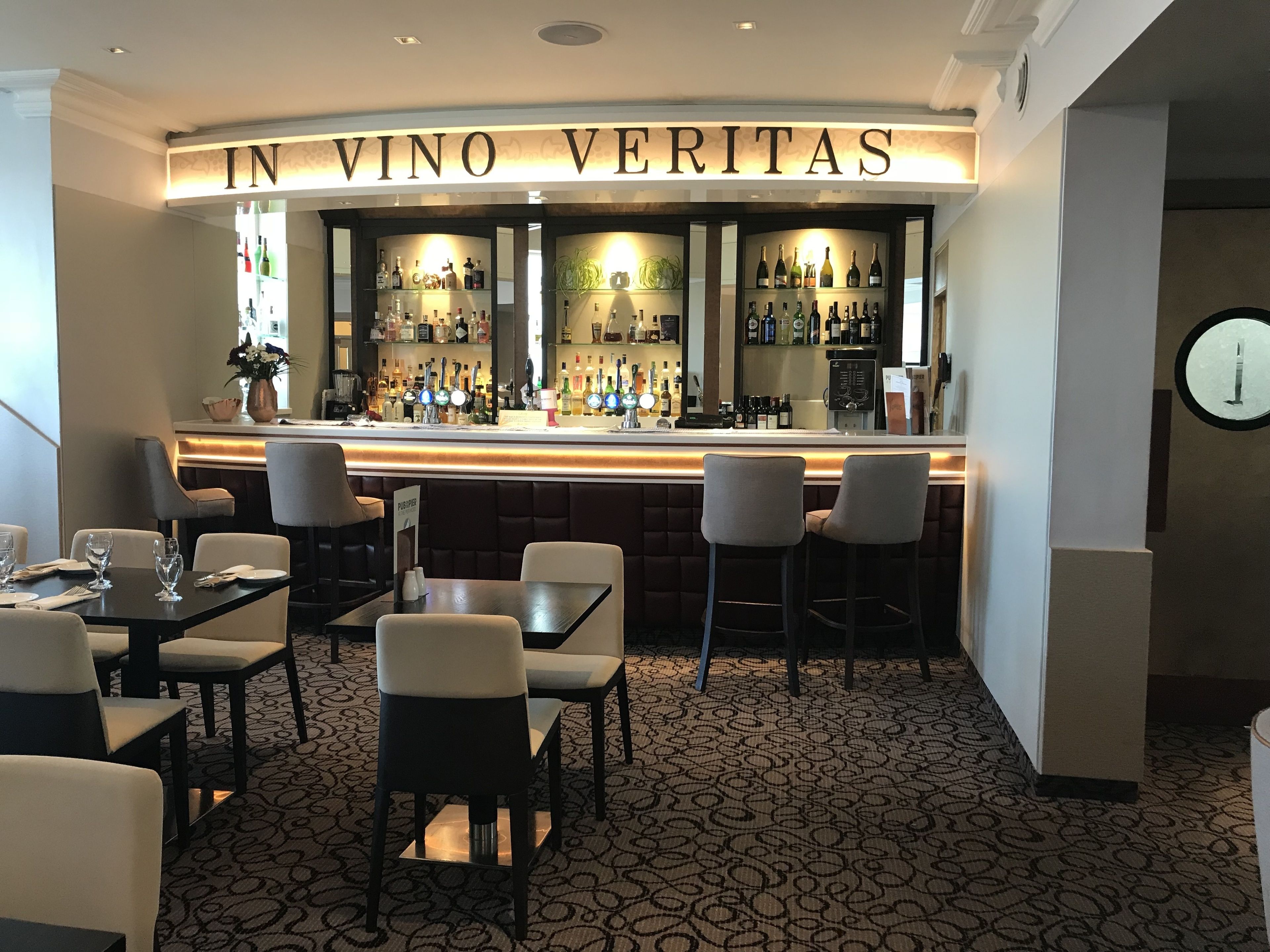 A bar area with a sign reading "IN VINO VERITAS" and a variety of bottles on shelves. There are several tables and chairs arranged for seating.
