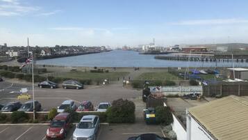 A parking lot with several cars, a river, and a distant view of a town with buildings and a dock.