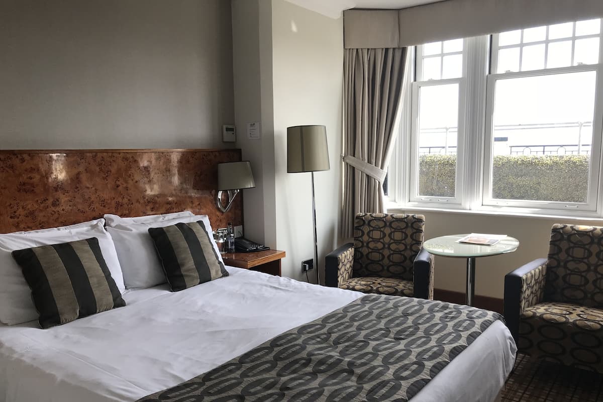Une chambre d’hôtel avec un lit, deux chaises, une petite table, une lampe et une fenêtre avec des rideaux.