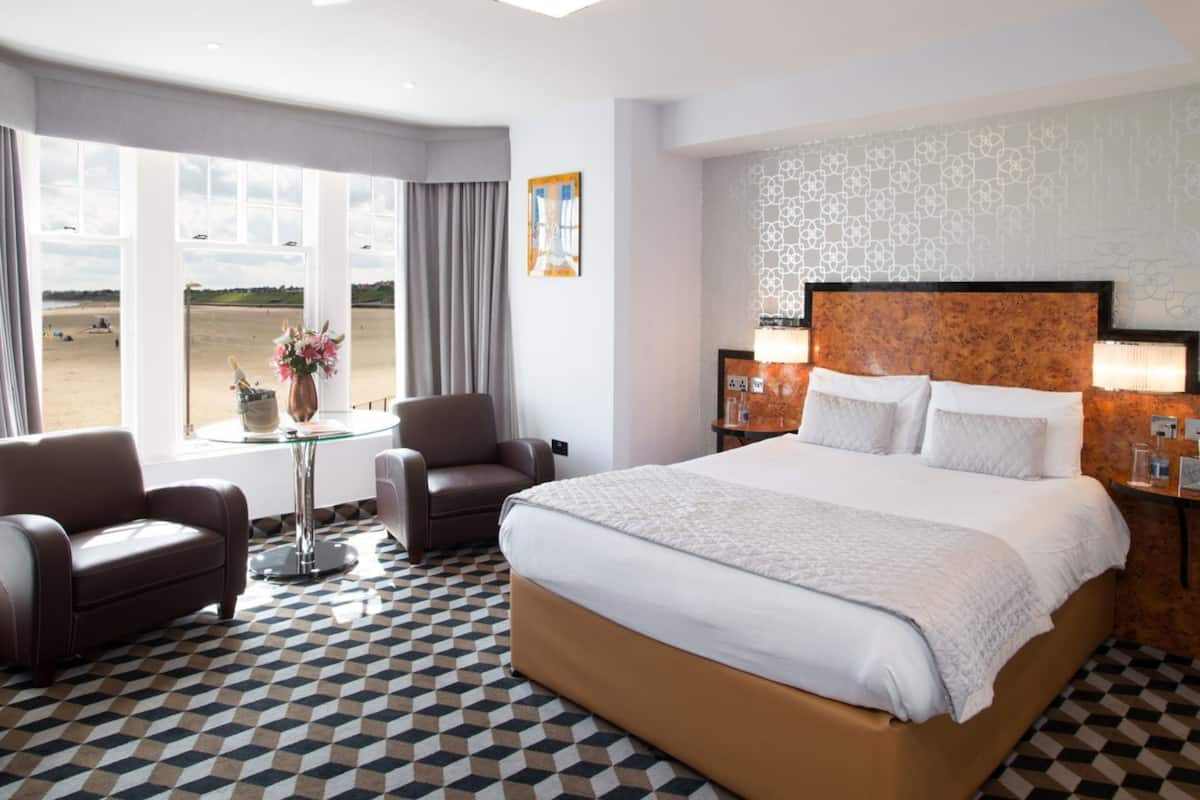 Une chambre d’hôtel avec un grand lit, deux fauteuils, une petite table sur laquelle est posé un vase de fleurs, une vue sur la plage et un sol à motifs.