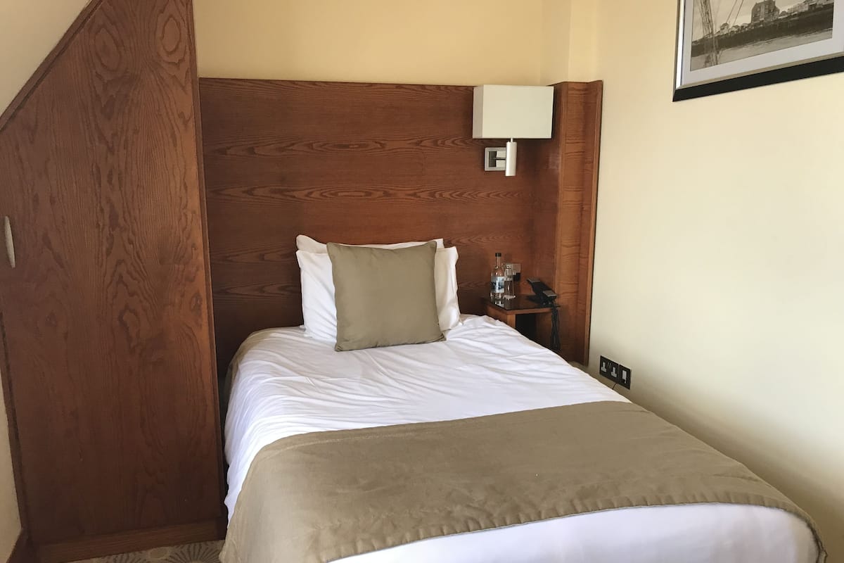 Une chambre d’hôtel avec une tête de lit en bois, un lit recouvert de draps blancs, un plaid beige, une table de chevet avec une lampe et un tableau encadré accroché au mur.