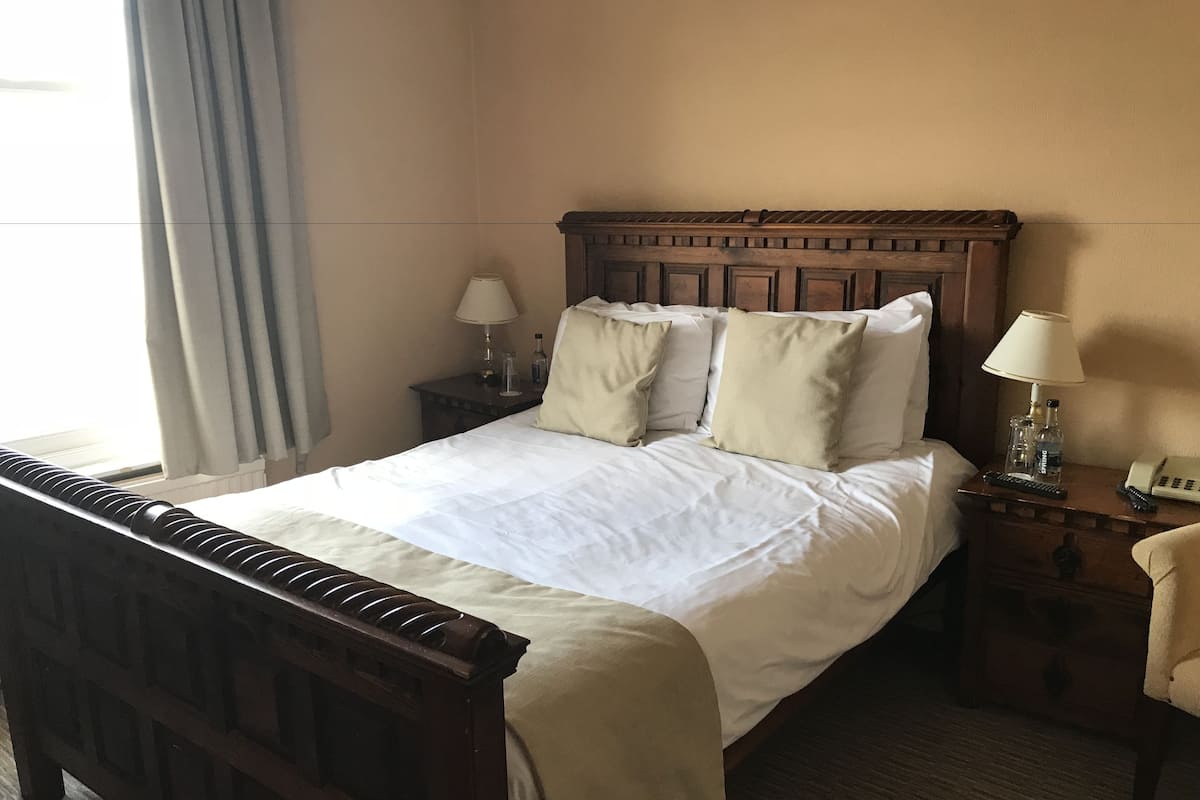 Une chambre d’hôtel avec un lit en bois, deux tables de chevet avec des lampes, une petite table avec un téléphone et une fenêtre avec des rideaux.