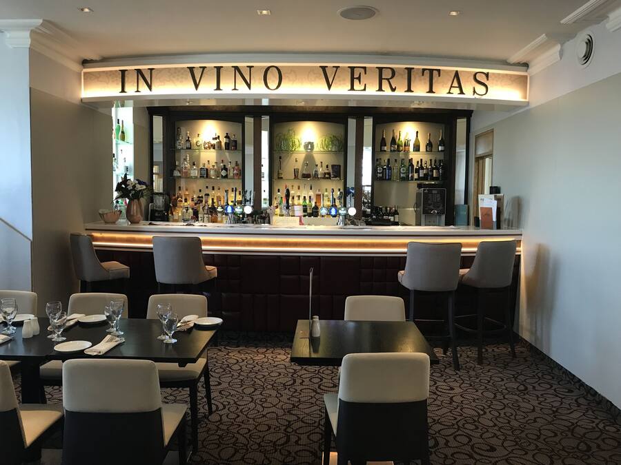 A bar area with a sign reading "IN VINO VERITAS" and a selection of bottles and glasses. There are tables and chairs arranged for seating.