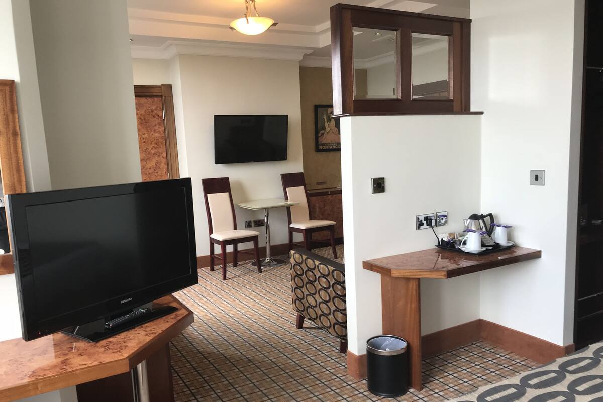 Une chambre d’hôtel équipée d’une télévision, d’une petite table à manger avec des chaises, d’un coin café et d’une poubelle.