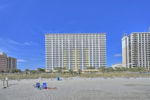 A high-rise hotel with multiple floors, a beach in the foreground, and palm trees.