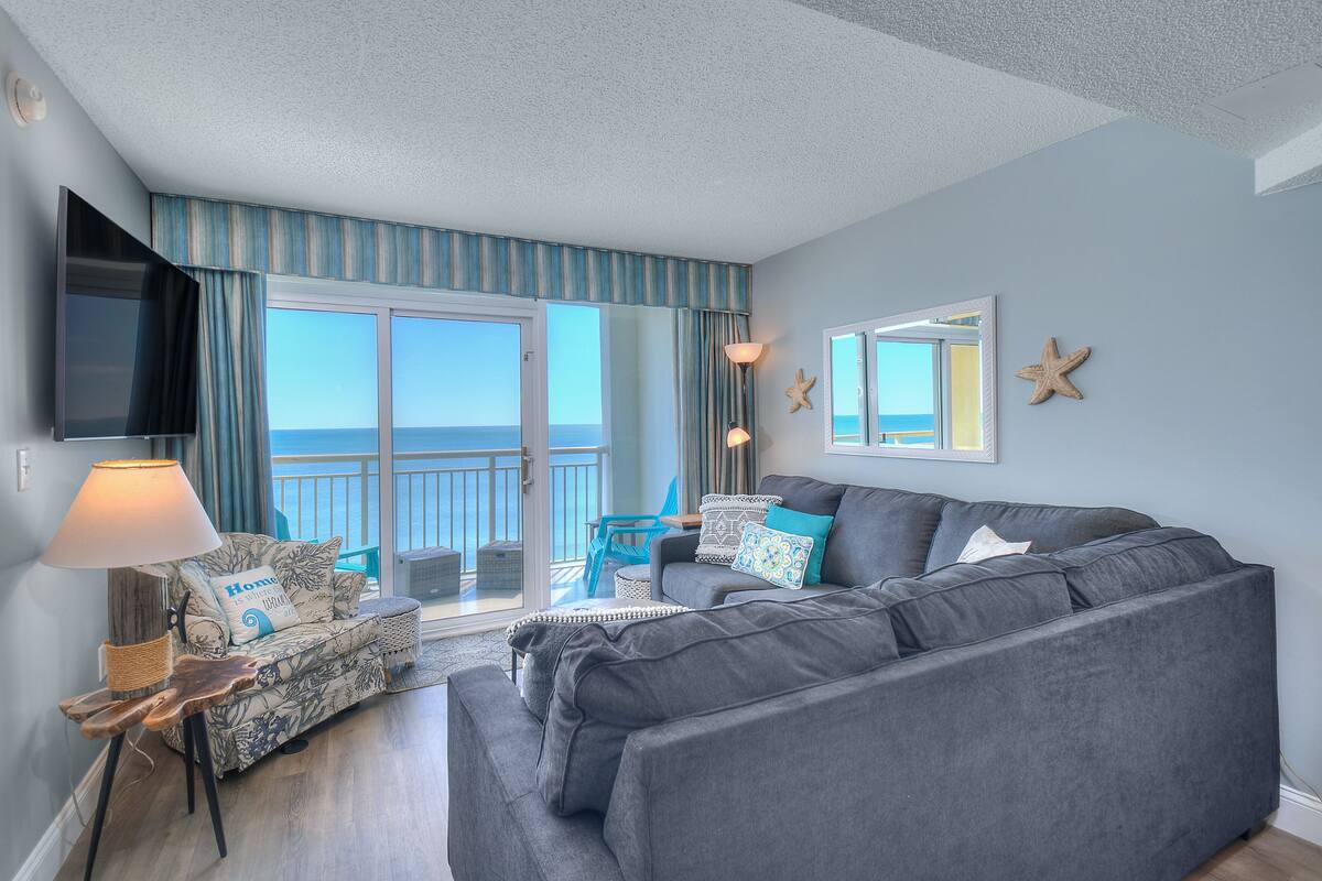 Un salon avec un canapé gris, un fauteuil bleu, un téléviseur et un balcon offrant une vue sur la mer.