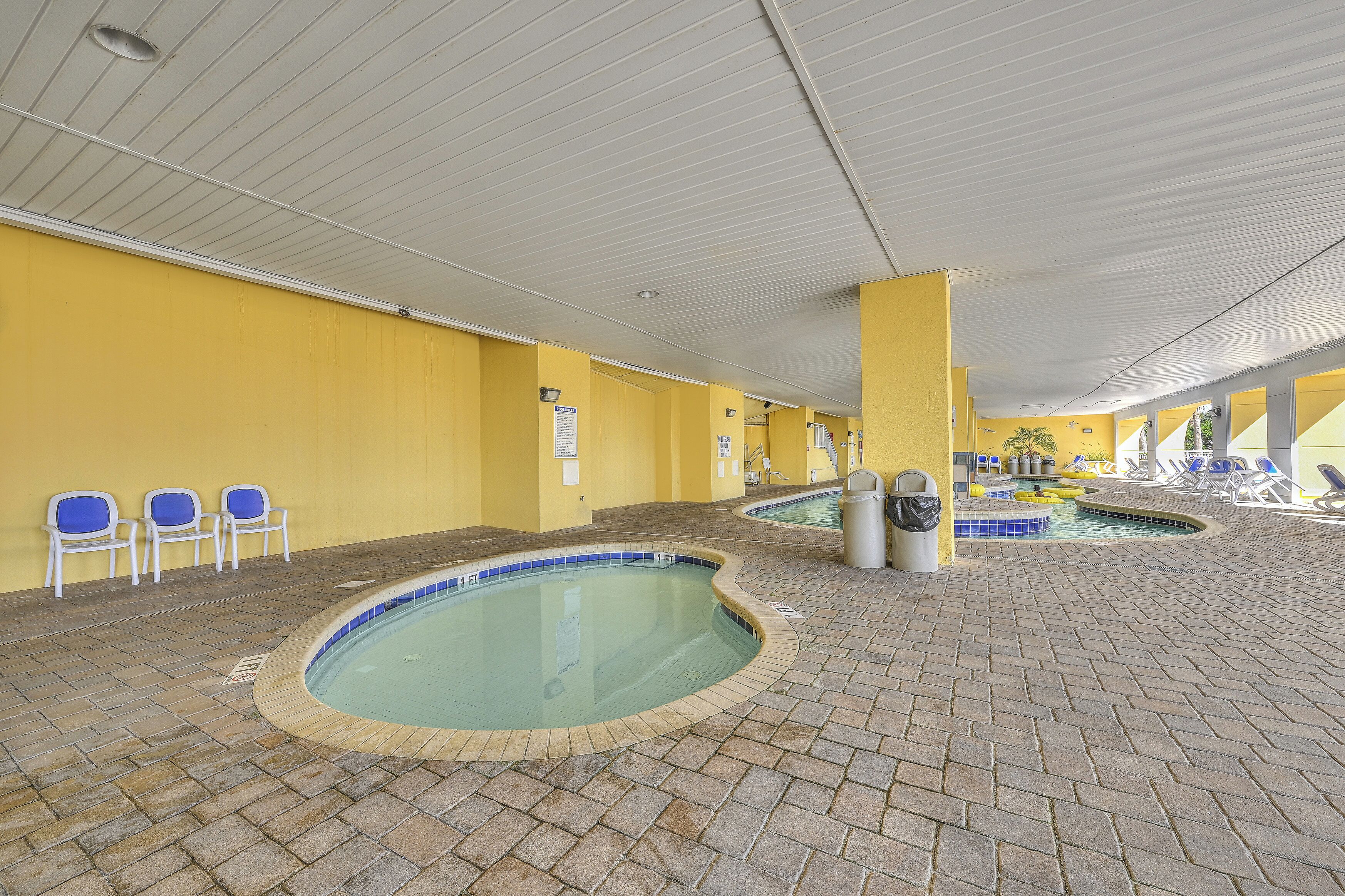 Un espace piscine intérieure comprenant plusieurs bassins, des sols carrelés et des chaises bleues se détachant sur des murs jaunes.