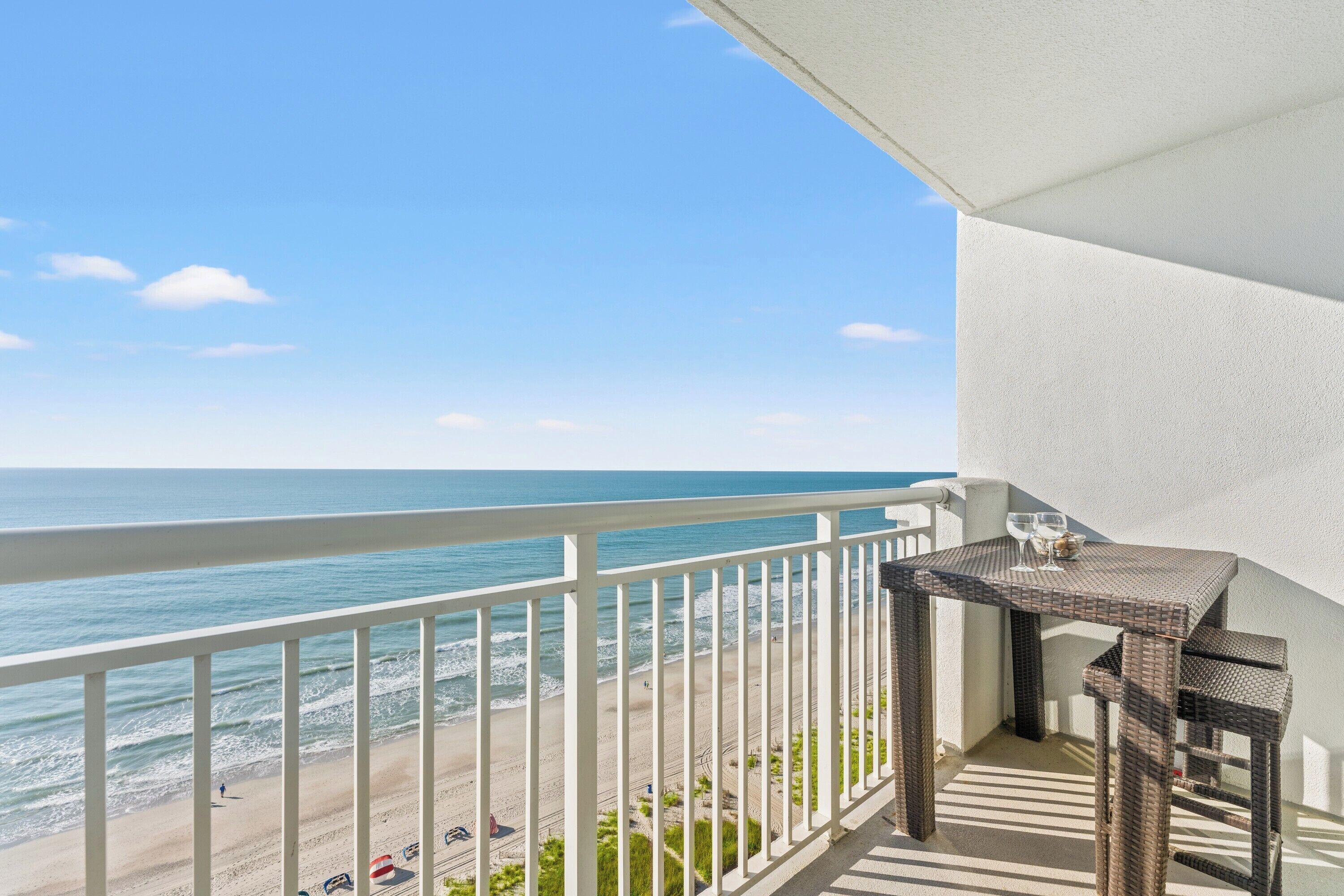 A balcony with a view of the beach and ocean, a wicker table with two chairs, and a white railing.