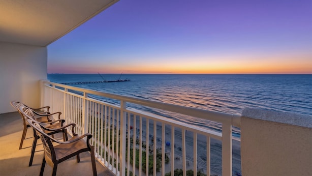 A balcony with a view of the sea at sunset, wooden chairs, and a white railing. - Camelot by the Sea by Elliott Beach Rentals (Myrtle Beach)