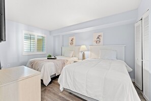 A bedroom with two beds, a bedside table, a lamp, and a window with blinds. - Camelot by the Sea by Elliott Beach Rentals (Myrtle Beach)
