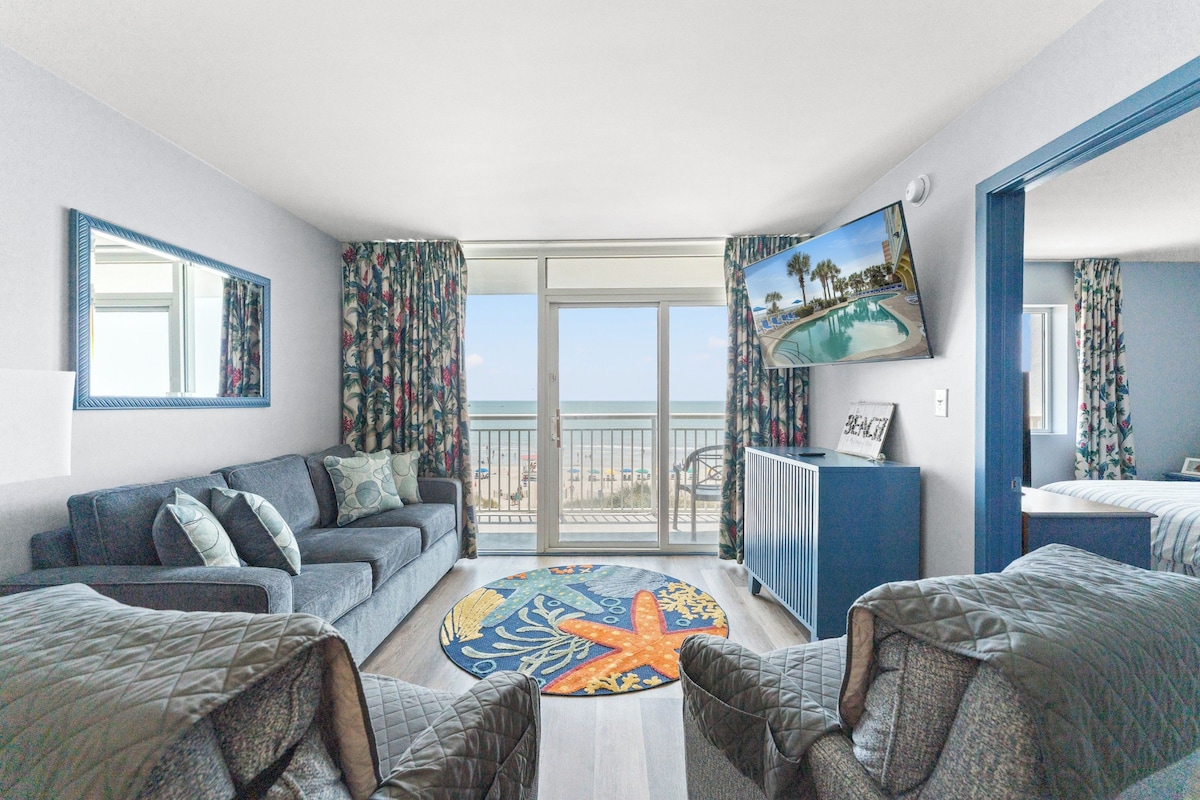 A hotel room with a balcony, a sofa, a chair, a small table, and a TV.