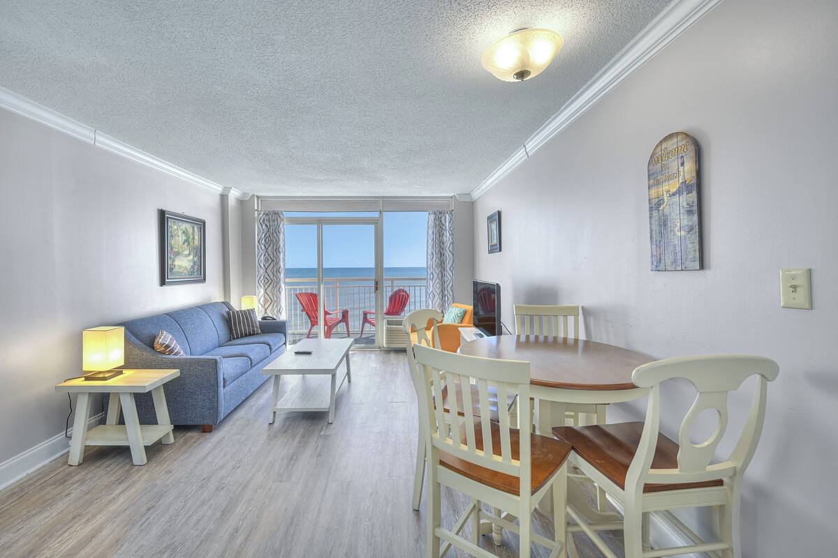 A living room with a sofa, coffee table, and dining table with chairs. There is a view of the ocean through large windows.