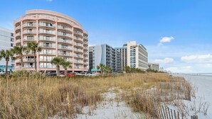 A beachfront hotel with multiple balconies, a sandy beach, and a clear sky. - Units at St. Clements at Caravelle by Elliott Beach Rentals (Myrtle Beach)