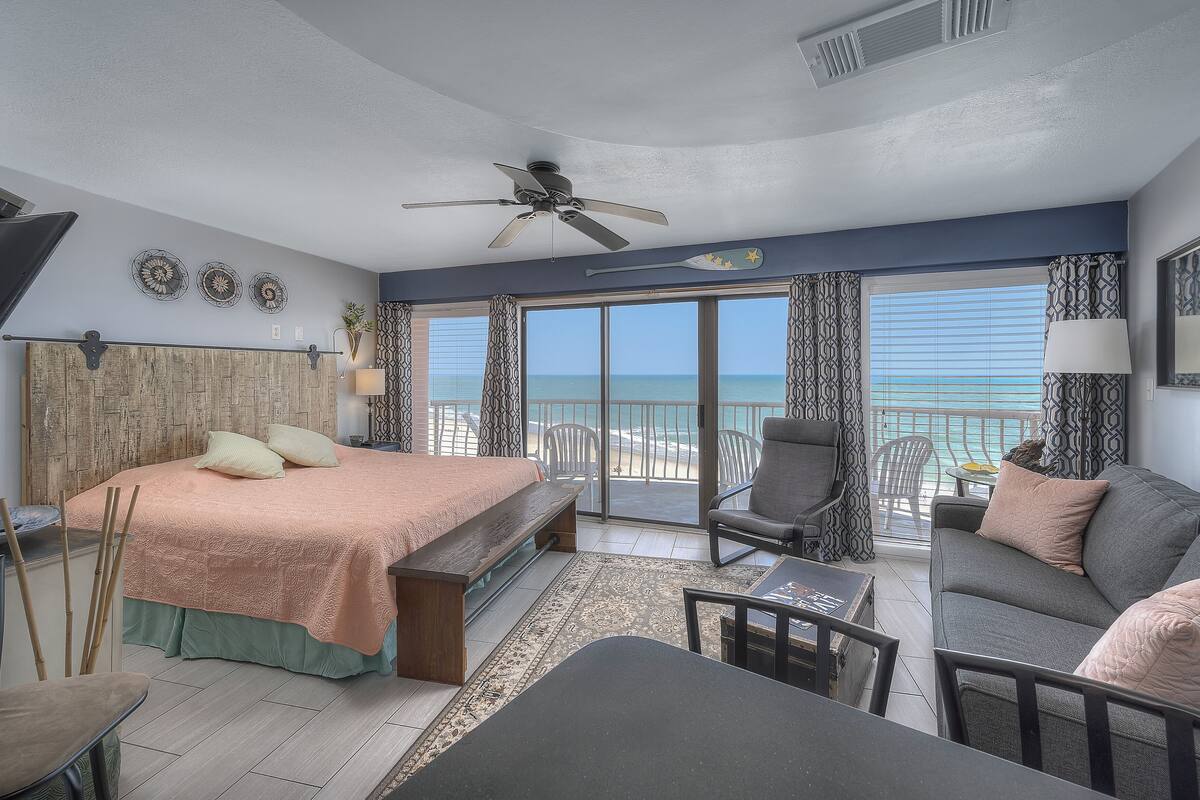 A bedroom with a bed, a sofa, a chair, a table, and a ceiling fan. There is a large window with a view of the ocean.