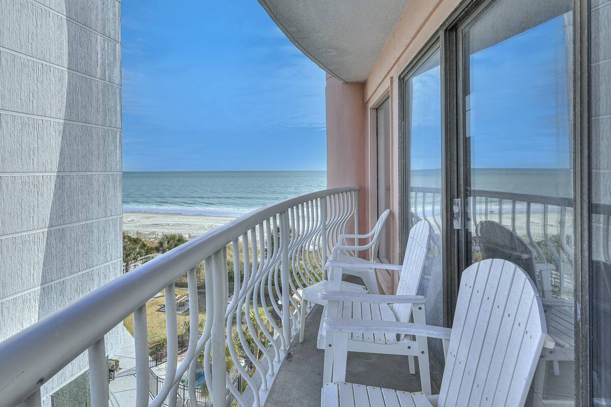 A balcony with white Adirondack chairs overlooking a beach.