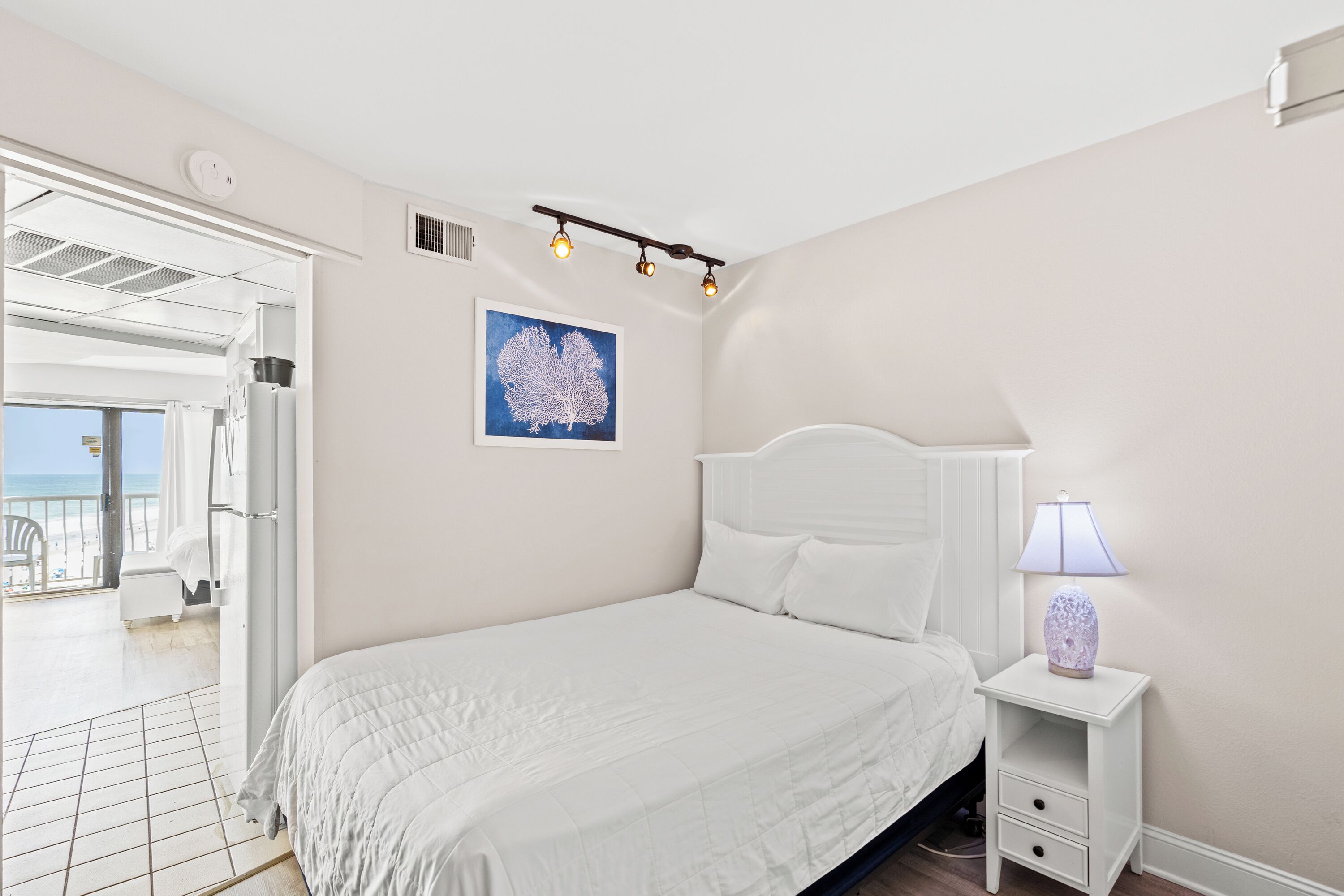 A bedroom with a bed, a nightstand with a lamp, a ceiling light fixture, and a framed artwork on the wall.