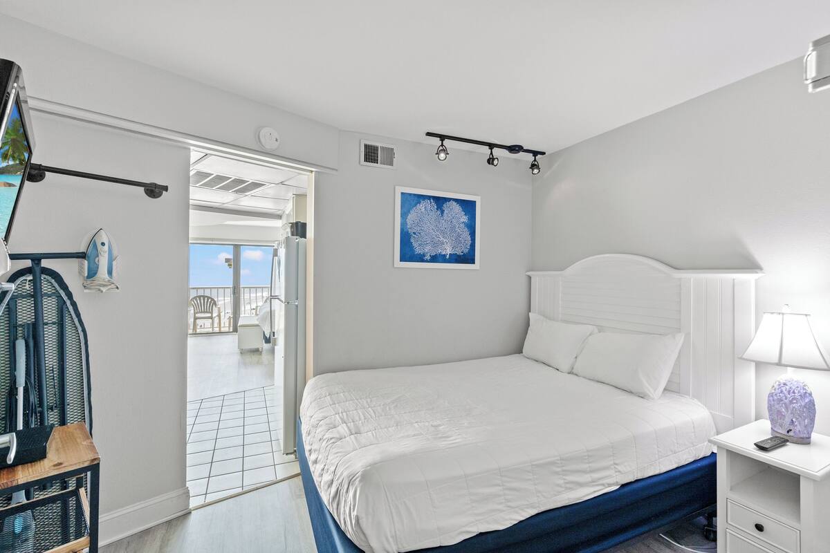 A bedroom with a bed, a nightstand, a lamp, a picture on the wall, and a view of the outside.