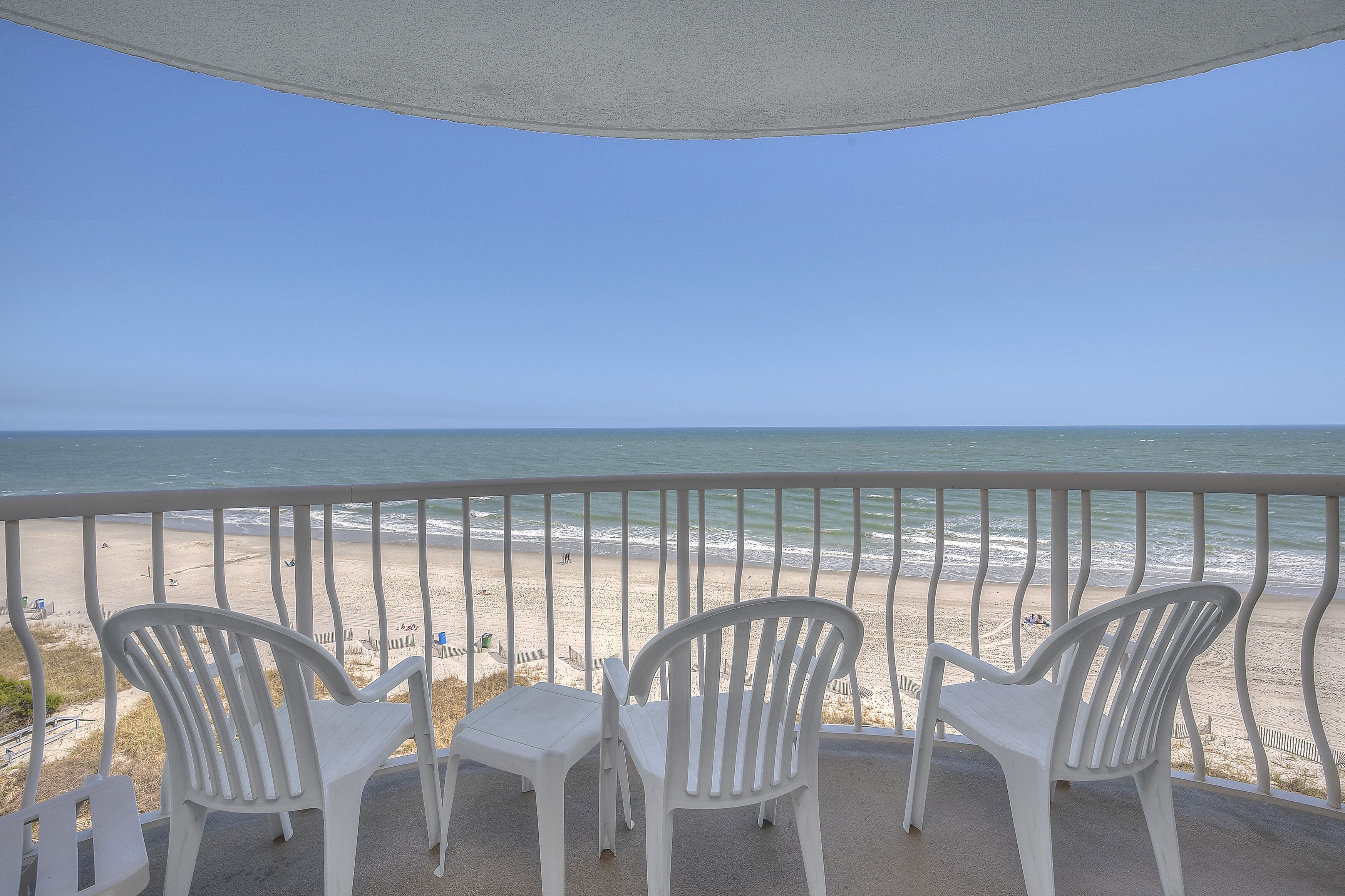 A balcony with white plastic chairs and a table overlooking a beach with a clear view of the ocean.