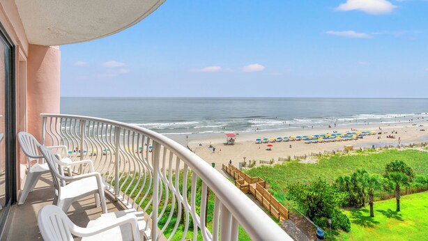 A balcony with white plastic chairs and a view of the beach with umbrellas and people. - Units at St. Clements at Caravelle by Elliott Beach Rentals (Myrtle Beach)