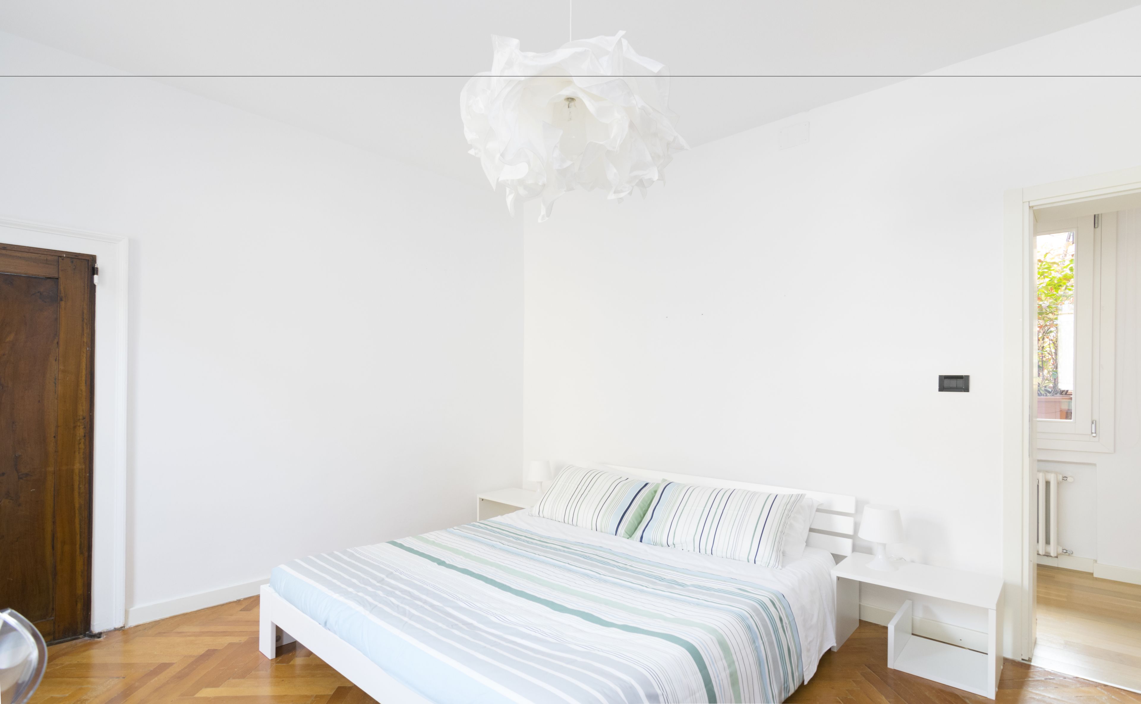 Une chambre à coucher avec un lit, une table de chevet blanche, un luminaire suspendu et une porte en bois.