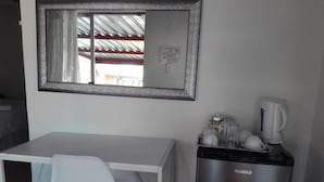 A compact hotel room with a white desk, a chair, a small refrigerator, and a coffee maker. - Joezebel Guest House (Moses Kotane)