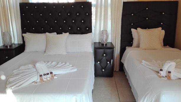 A hotel room with two beds, each with a black tufted headboard and white bedding. There are two nightstands with lamps, a small table with a vase, and a towel rack with towels and toiletries. - Joezebel Guest House (Moses Kotane)