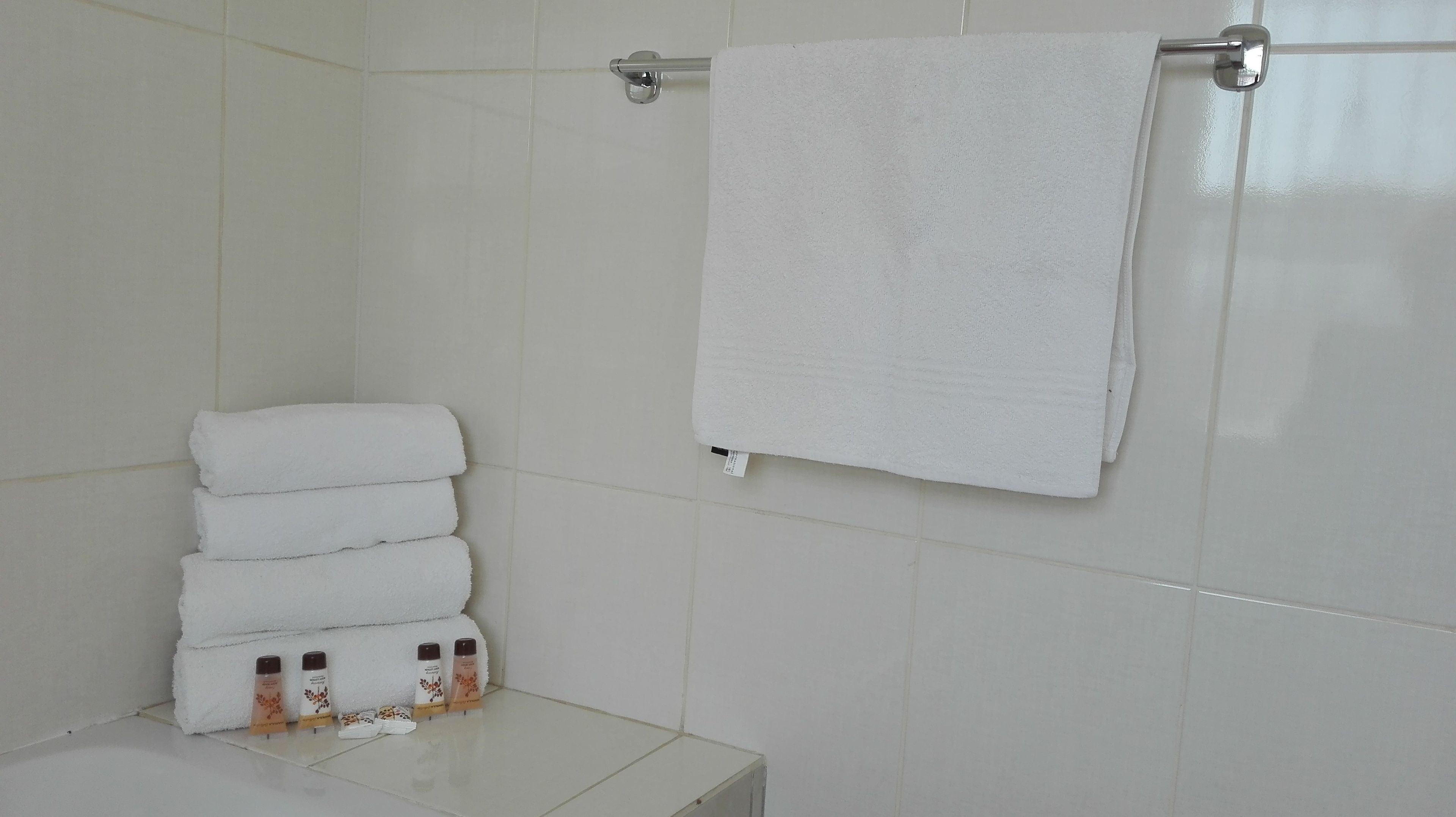 A bathroom with white tiled walls, a white towel rack with a folded towel, and a stack of white towels.