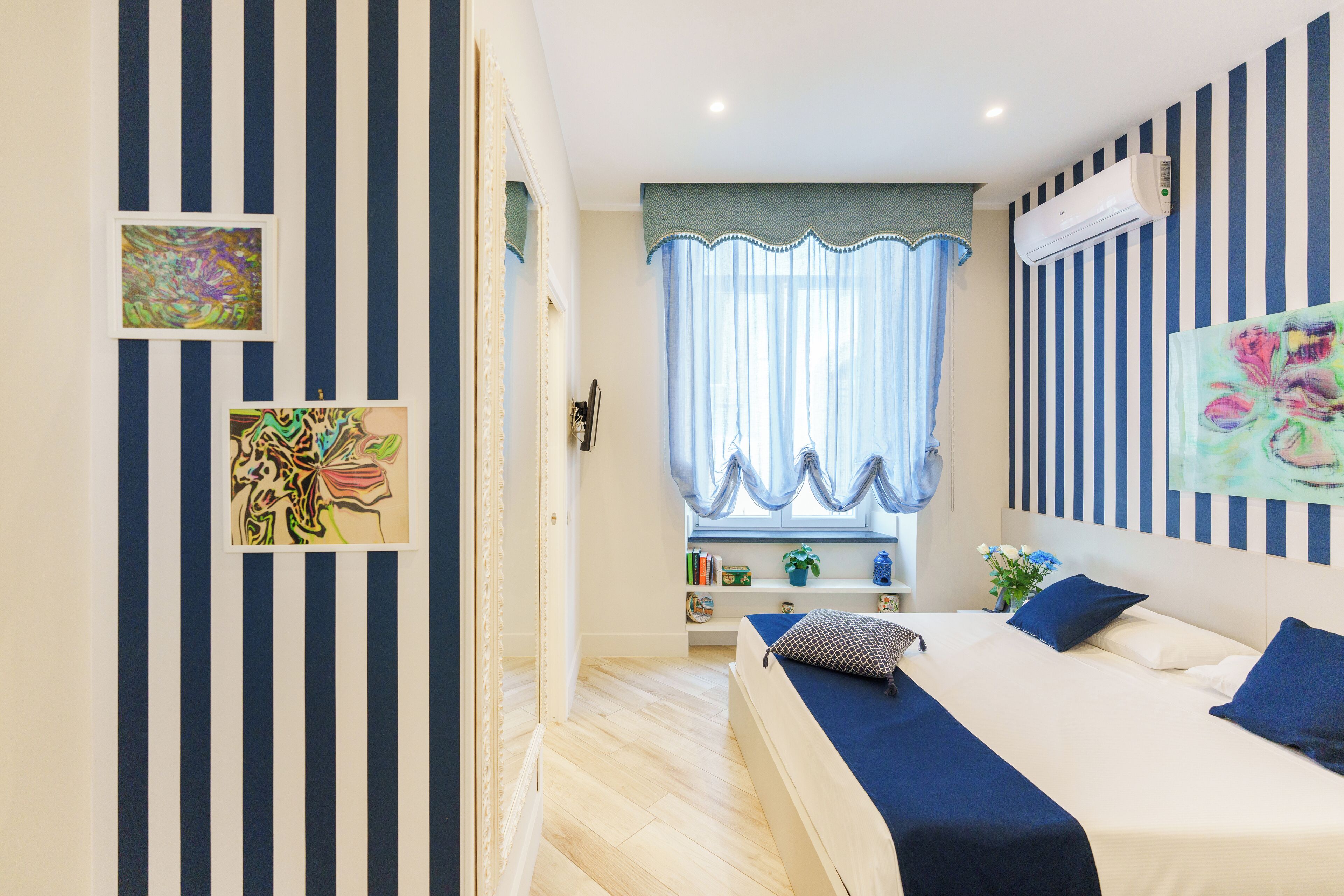 A bedroom with striped walls, a bed with blue bedding, a window with curtains, and artwork on the walls.