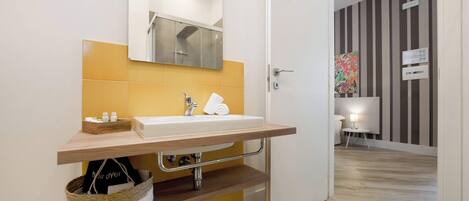 A modern bathroom with a wooden vanity, a mirror, and a sink. There is a basket under the vanity and a towel hanging on the wall.