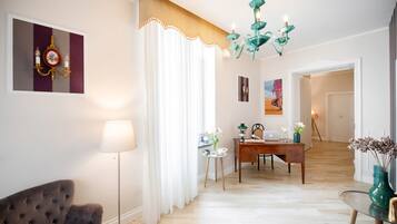 A well-lit living room with a sofa, a lamp, a dining table with chairs, a chandelier, and a painting on the wall.
