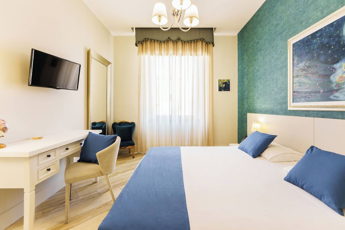 Ein Hotelzimmer mit Bett, Schreibtisch, Stuhl, Fernseher und einem Gemälde an der Wand.