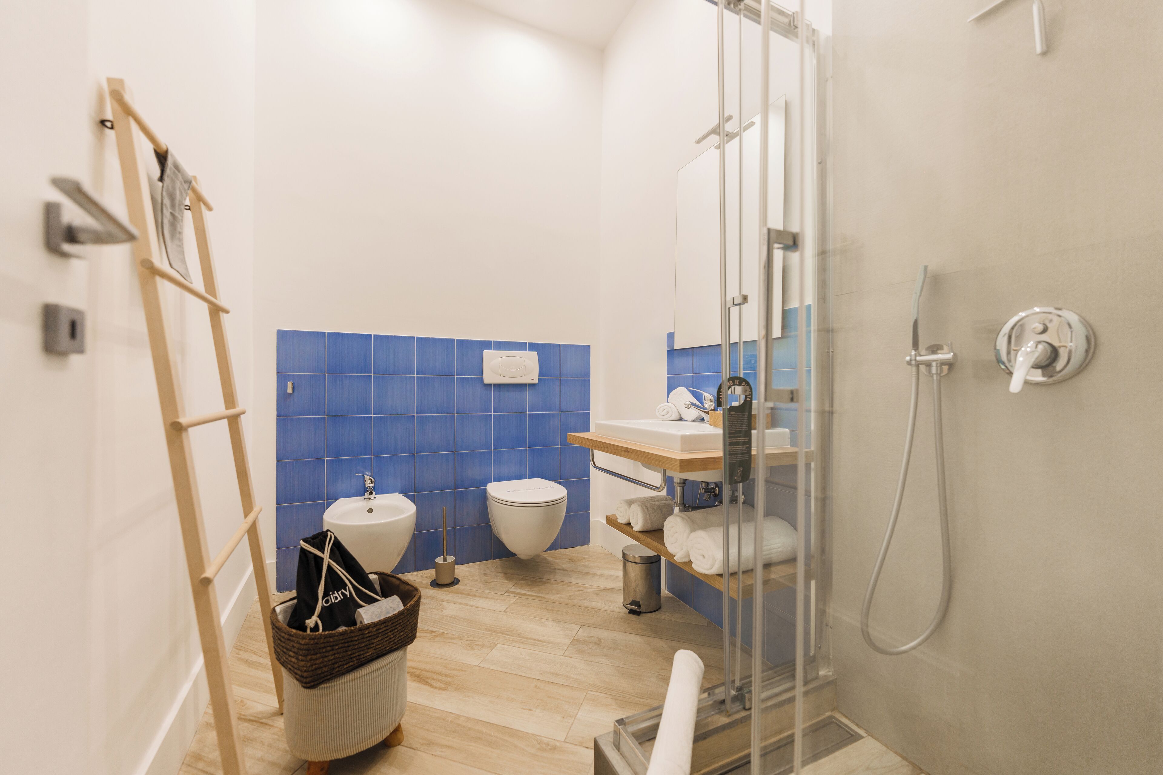 Une salle de bain moderne avec une douche à parois de verre, une échelle en bois, un lavabo blanc et un mur recouvert de carreaux bleus.
