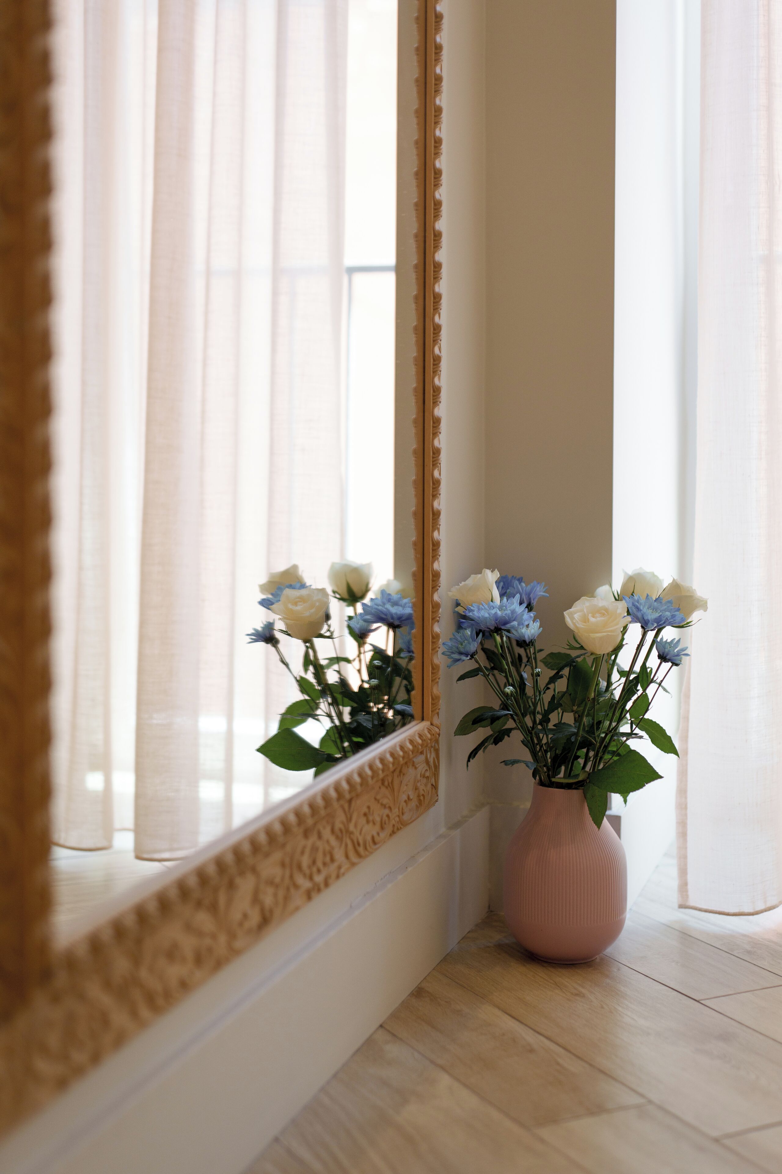 Eine Ecke mit Spiegel, der ein Fenster spiegelt, einer rosa Vase mit Blumen und einem Holzboden.