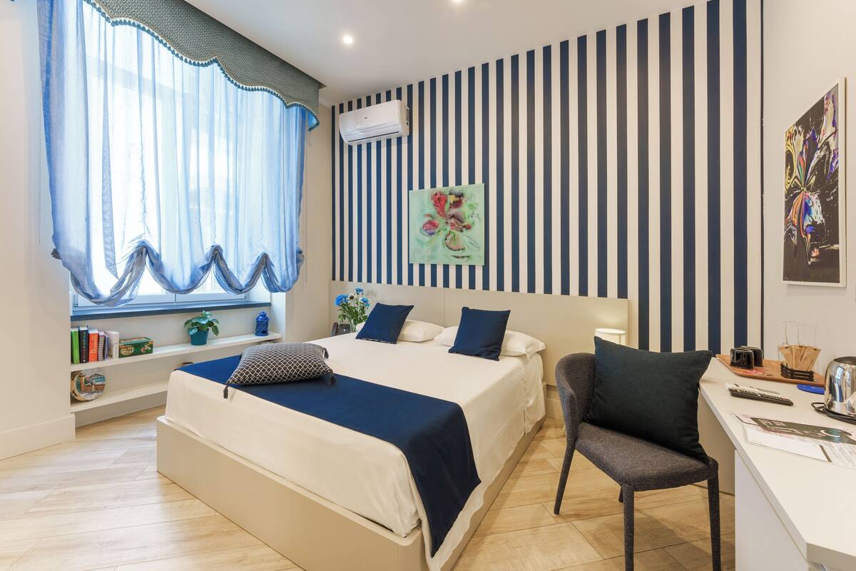 Ein modernes Schlafzimmer mit einem großen Bett, einem Schreibtisch und einem Stuhl. Ein Fenster mit transparenten Vorhängen und ein Blumentopf auf der Fensterbank.