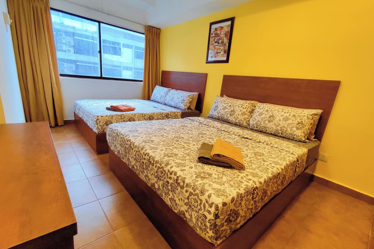 A hotel room with two beds, a wooden headboard, a large window with curtains, a framed picture on the wall, and a wooden table.