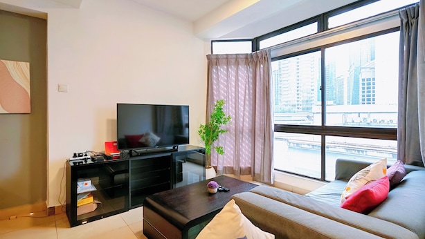 A modern living room with a flat-screen TV, a sofa with cushions, a coffee table, and a large window with a view of buildings. - Pavilion Fahrenheit 88 - AJ home (Kuala Lumpur)