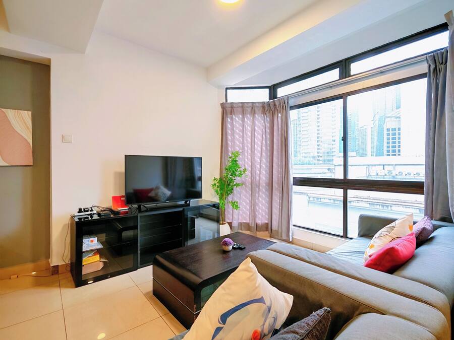 Uma sala de estar moderna com televisão de tela plana, um sofá com almofadas, uma mesa de centro e uma janela grande com vista para edifícios.