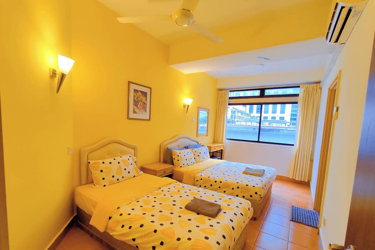 A hotel room with two beds, a ceiling fan, a window with a view of buildings, and a door with a handle.