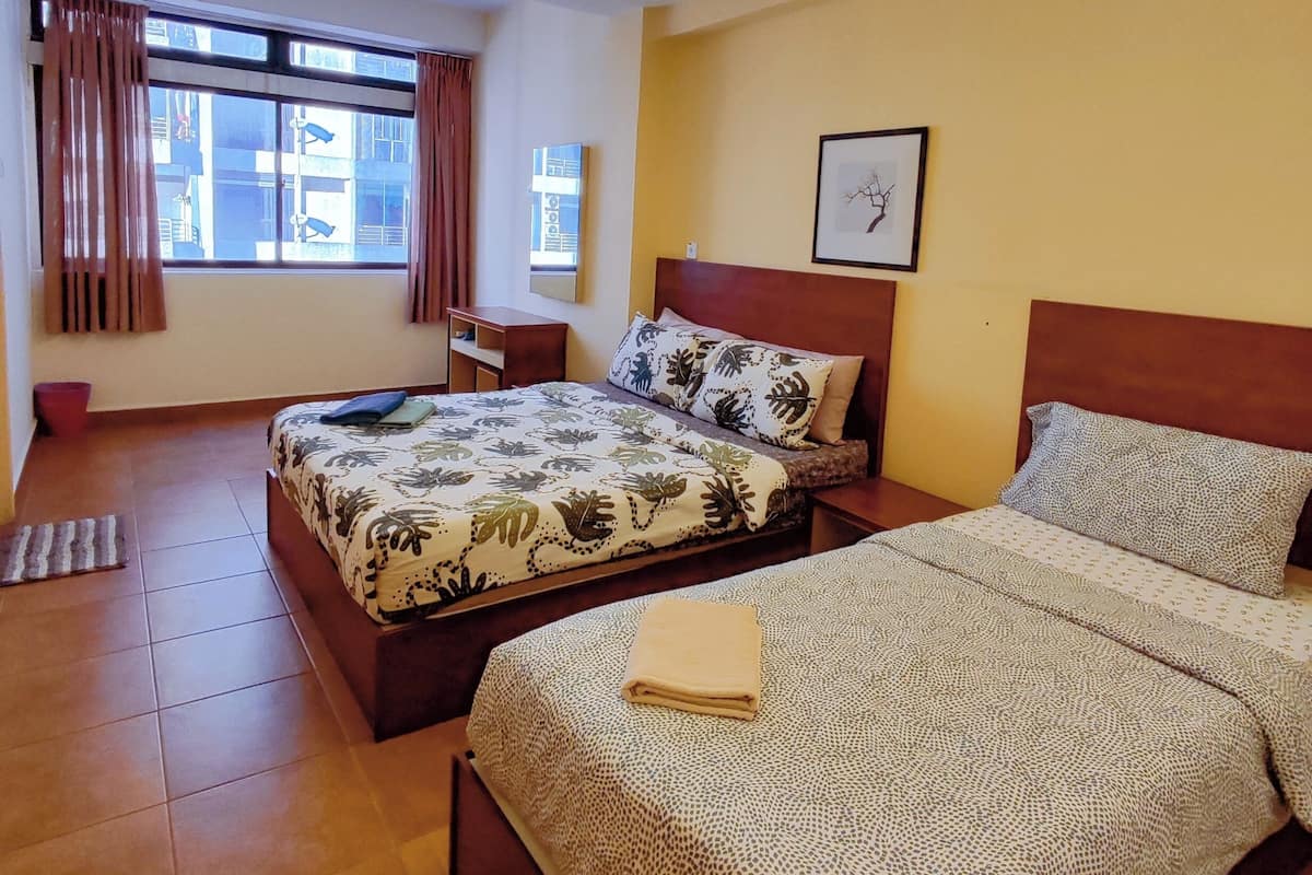 A hotel room with two beds, a ceiling fan, a window with curtains, a bedside table, a wall-mounted TV, and a framed picture.